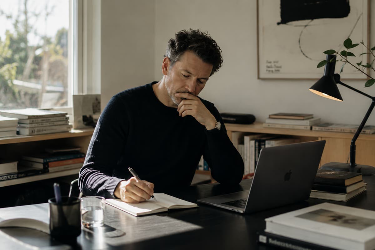 A founder thinking over a notebook at a desk, pen in hand and a laptop open to one side.