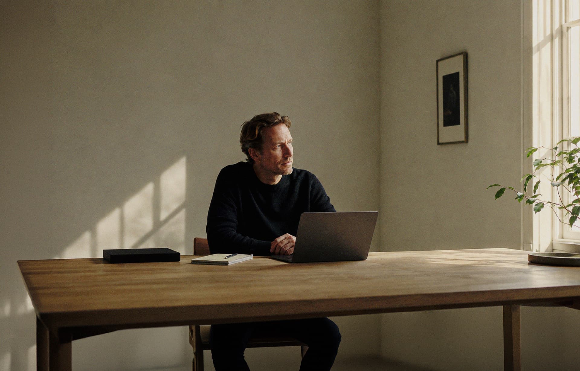A writer at a long wooden table in a quiet, sunlit room, a laptop and a closed notebook in front of him, looking out of the window.